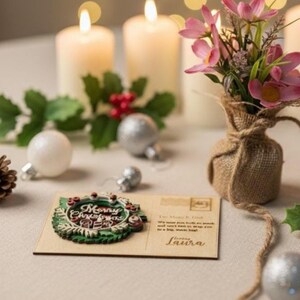May include: A Christmas-themed scene with lit candles, ornaments, and a postcard. The postcard features a wreath design with the words "Merry Christmas". A small burlap vase holds pink flowers, adding a touch of color to the festive arrangement.