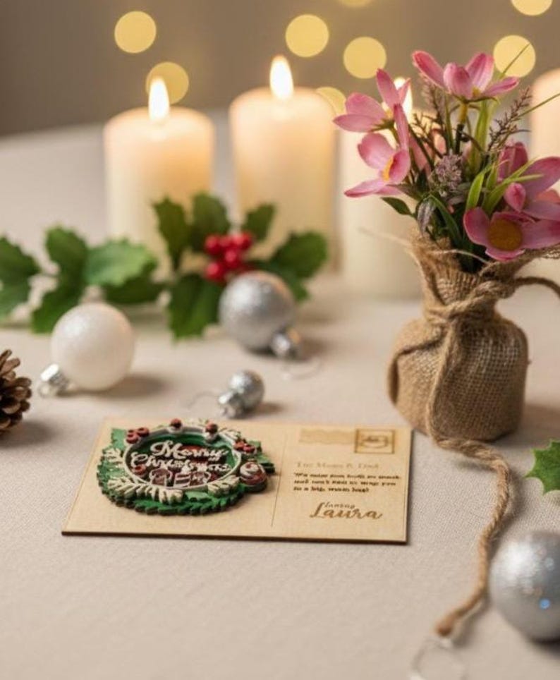 May include: A Christmas-themed scene with lit candles, ornaments, and a postcard. The postcard features a wreath design with the words "Merry Christmas". A small burlap vase holds pink flowers, adding a touch of color to the festive arrangement.