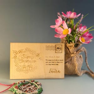 May include: A wooden postcard with the words "Merry Christmas" engraved in a wreath design. The postcard has a stamp and a message to Mom & Dad. A Christmas ornament and a small vase of pink flowers are also in the image.