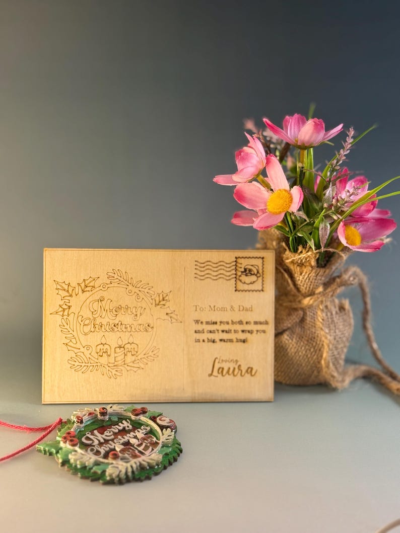 May include: A wooden postcard with the words "Merry Christmas" engraved in a wreath design. The postcard has a stamp and a message to Mom & Dad. A Christmas ornament and a small vase of pink flowers are also in the image.