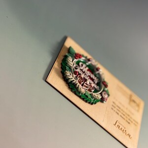 May include: A wooden postcard with a 3D Christmas wreath design. The wreath features green leaves, red berries, and the words "Merry Christmas" in the center. The postcard is addressed to "Laura".