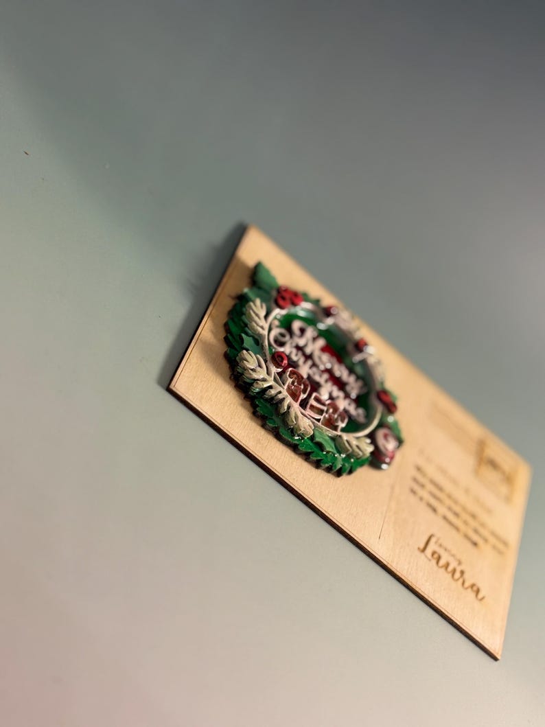 May include: A wooden postcard with a 3D Christmas wreath design. The wreath features green leaves, red berries, and the words "Merry Christmas" in the center. The postcard is addressed to "Laura".