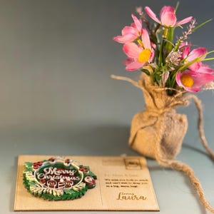 May include: A wooden Christmas card with a green and red wreath design that says "Merry Christmas". The card has a message to "Mom & Dad" and is signed "Loving Laura". A small bouquet of pink flowers sits in a burlap sack.