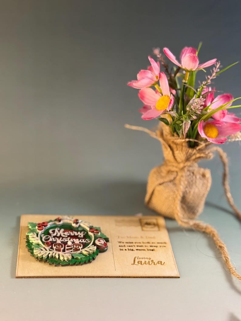 May include: A wooden Christmas card with a green and red wreath design that says "Merry Christmas". The card has a message to "Mom & Dad" and is signed "Loving Laura". A small bouquet of pink flowers sits in a burlap sack.