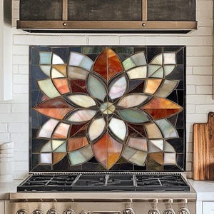 May include: A colorful stained glass backsplash with a floral design above a stainless steel stove. The flower design features shades of orange, red, green, and white. The kitchen has white cabinets and a dark range hood.