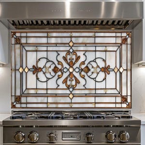 May include: A decorative stained glass backsplash above a stainless steel gas range. The glass features a geometric design with brown, gold, and white elements. The kitchen has white cabinets and a stainless steel range hood.
