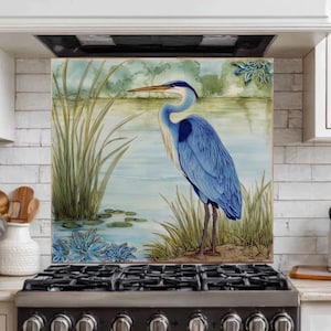 May include: A blue heron stands in a serene waterscape, depicted on a kitchen backsplash. The artwork features the bird with detailed blue plumage, set against a backdrop of water, reeds, and foliage. The backsplash is installed above a black stove.
