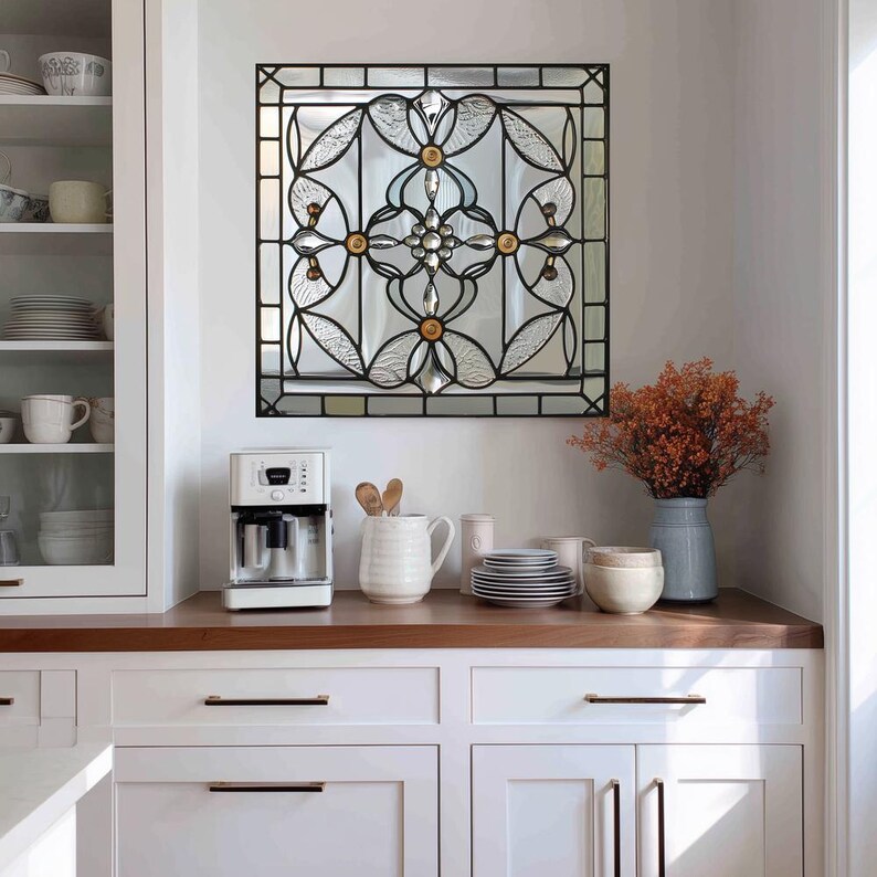 May include: A square stained glass window with a black metal frame is mounted on a white wall. The window features a symmetrical floral design with clear and amber-colored glass. A white coffee maker and kitchenware are on the countertop.