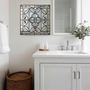 May include: Square, stained glass mirror with a black metal frame. The mirror features a floral design with clear and gold-toned accents. The mirror is mounted on a white wall in a bathroom setting.