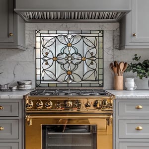 May include: A kitchen with a gold-colored stove and a decorative stained glass panel. The glass panel features a floral design with gold and clear glass elements. Gray cabinets and a marble countertop complete the kitchen's design.