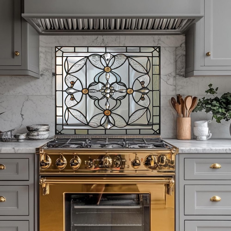 May include: A kitchen with a gold-colored stove and a decorative stained glass panel. The glass panel features a floral design with gold and clear glass elements. Gray cabinets and a marble countertop complete the kitchen's design.