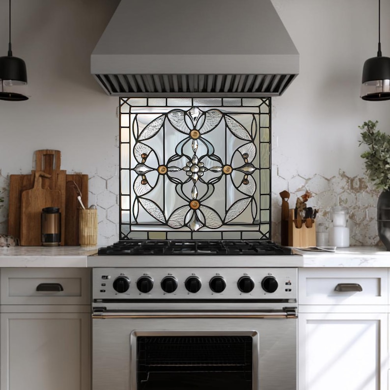 May include: A decorative stained glass panel with a floral design is installed above a stainless steel range. The glass features clear and amber-colored elements within a black metal frame. The kitchen setting includes white cabinetry and a stainless steel range hood.