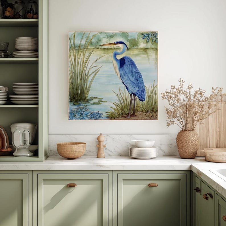 May include: A framed watercolor painting of a blue heron standing near a lake. The artwork features shades of blue, green, and brown, with tall grasses and water lilies in the foreground. The heron has a long beak and stands tall.