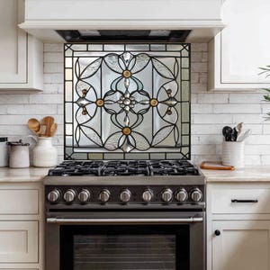 May include: A decorative stained glass panel with a black metal frame, featuring a floral design with clear and textured glass, and gold-colored accents. The panel is installed above a stainless steel gas range in a kitchen setting.