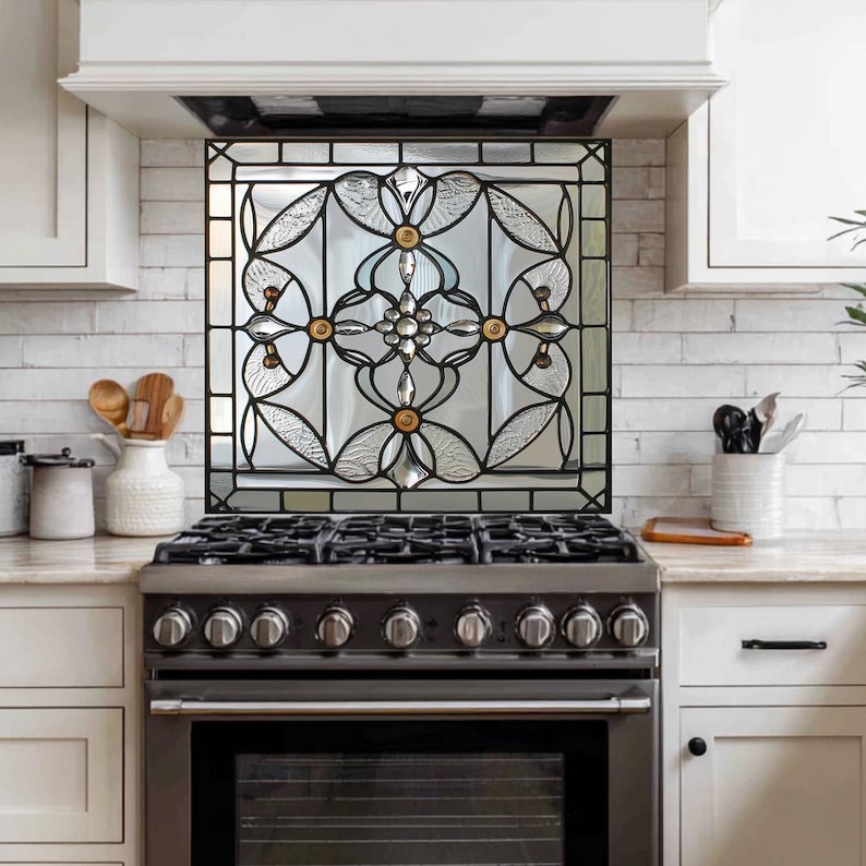 May include: A decorative stained glass panel with a black metal frame, featuring a floral design with clear and textured glass, and gold-colored accents. The panel is installed above a stainless steel gas range in a kitchen setting.