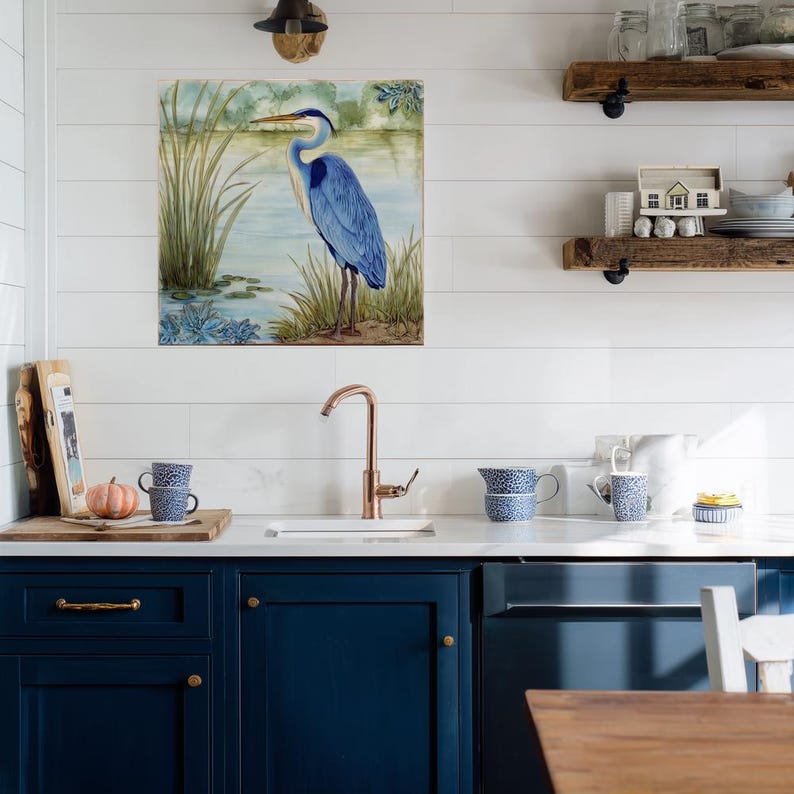 May include: A blue heron art print hangs above a kitchen sink. The artwork depicts a blue heron standing in a marshy area with reeds and water. The kitchen features navy blue cabinets, a copper faucet, and rustic wooden shelves.