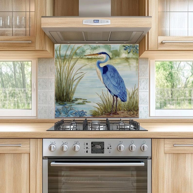 May include: A decorative kitchen backsplash featuring a blue heron standing near a body of water with reeds and lily pads. The artwork is framed by a light blue and green background, creating a serene, nature-inspired scene. The backsplash is installed above a gas stove.