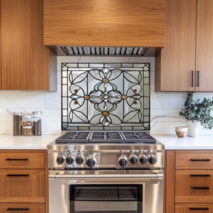 May include: A decorative stained glass backsplash with a floral design is installed above a stainless steel gas range. The glass features clear, amber, and black accents. The kitchen has wooden cabinets and a white countertop.