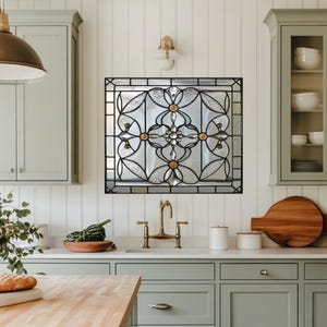 May include: A square stained glass window with a black metal frame, featuring a floral design with clear and frosted glass. The window is mounted on a white wall in a kitchen setting. The design includes gold and clear accents.
