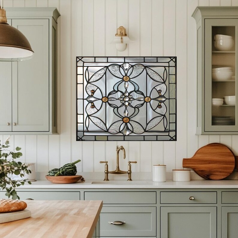 May include: A square stained glass window with a black metal frame, featuring a floral design with clear and frosted glass. The window is mounted on a white wall in a kitchen setting. The design includes gold and clear accents.