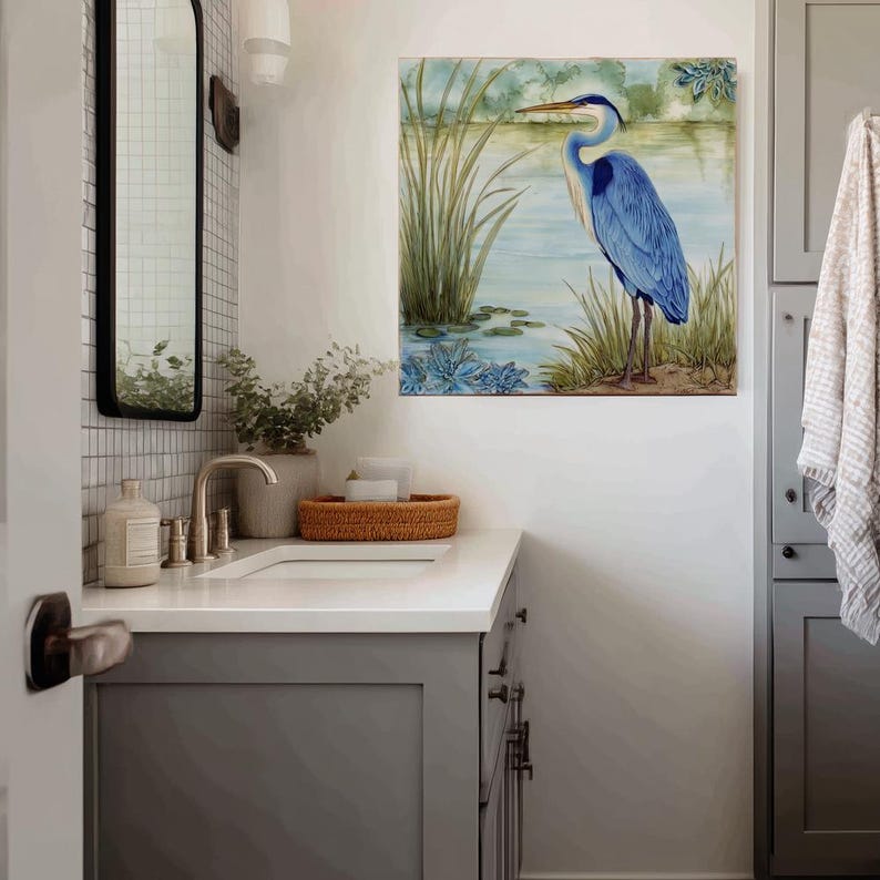 May include: A square wall art print featuring a blue heron standing in a marshy area with tall green reeds and blue water. The artwork has a watercolor style with soft colors and is displayed in a bathroom setting.