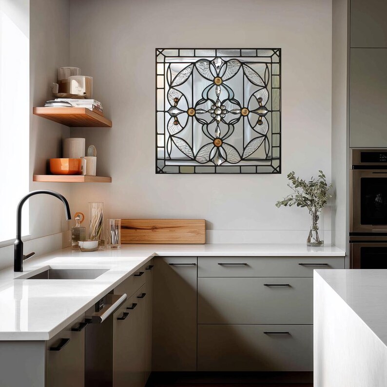 May include: A square stained glass window with a black metal frame is mounted on a wall. The glass features a floral design with clear and amber-colored elements. The kitchen has gray cabinets and a white countertop.
