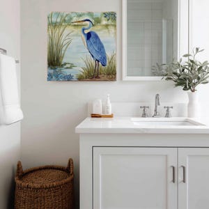 May include: A square watercolor painting of a blue heron standing in a marshy area. The artwork is displayed above a white bathroom vanity with a sink, mirror, and decorative items. A woven basket sits on the floor.