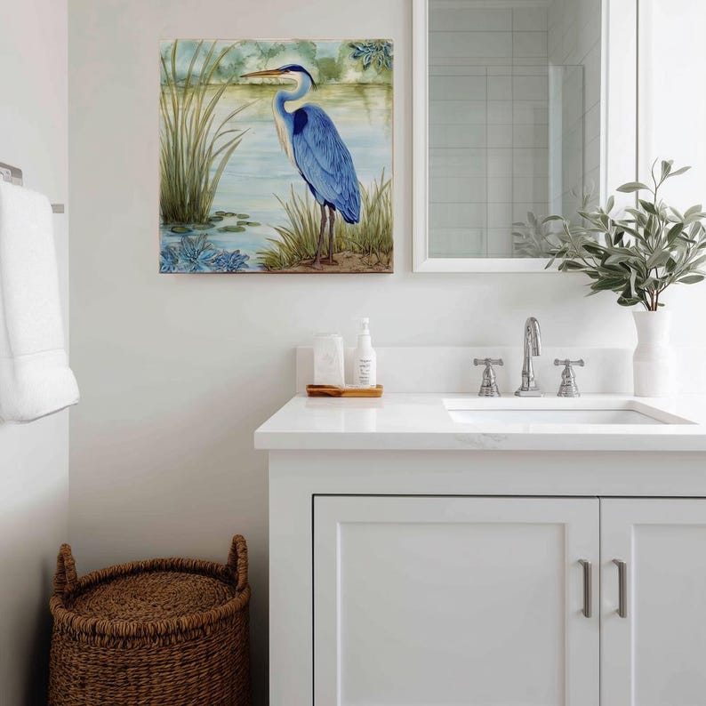 May include: A square watercolor painting of a blue heron standing in a marshy area. The artwork is displayed above a white bathroom vanity with a sink, mirror, and decorative items. A woven basket sits on the floor.