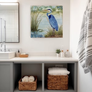 May include: A bathroom scene featuring a framed painting of a blue heron standing near a lake. The room includes a white countertop with a sink, a mirror, and woven baskets for storage. A gray and white striped towel hangs on the wall.