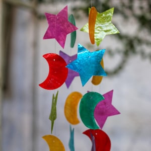 Puede incluir: Un móvil colorido con estrellas y medias lunas hechas de concha iridiscente. El móvil está suspendido de un anillo de madera y cuelga de una cuerda blanca.
