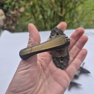 Puede incluir: Una manija de puerta de latón vintage con un diseño ornamentado. La manija tiene la forma de un rectángulo largo y delgado con un extremo curvo. La manija se sostiene en una mano.