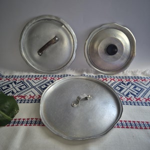 Puede incluir: Tres tapas de olla de aluminio plateado con asas. Las tapas son redondas y tienen un borde ligeramente elevado. Las asas son de metal y están unidas a las tapas con remaches.