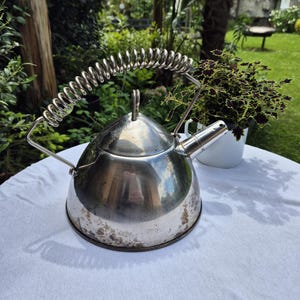 May include: A stainless steel tea kettle with a spring handle. The kettle has a conical shape and a spout. It sits on a white tablecloth outdoors, with a garden in the background. The kettle is approximately 8 inches in diameter.