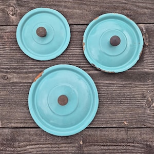 May include: Three vintage turquoise enamel pot lids with brown wooden knobs. The lids have a distressed, rustic appearance, with visible wear and tear around the edges. They are arranged on a weathered wooden surface, creating a charming, antique aesthetic.