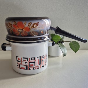Op de afbeelding: Een witte emaille dubbele kookpan met een zwarte handgreep en een bloemenontwerp aan de bovenkant. De bodem van de pan heeft een geometrisch patroon in zwart en rood.