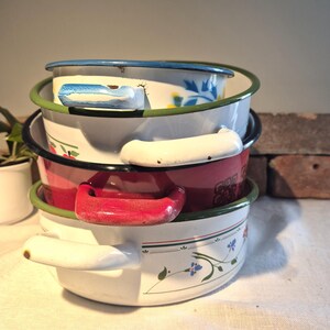 Può includere: Una pila di quattro pentole smaltate con motivi floreali. La pentola superiore è blu con fiori bianchi, la seconda è bianca con fiori rossi, la terza è rossa con motivi in ​​bianco e nero e la pentola inferiore è bianca con fiori verdi e blu.