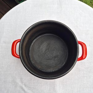 May include: A red enameled cast iron pot with black interior and two red handles. The pot is empty and sitting on a white tablecloth.