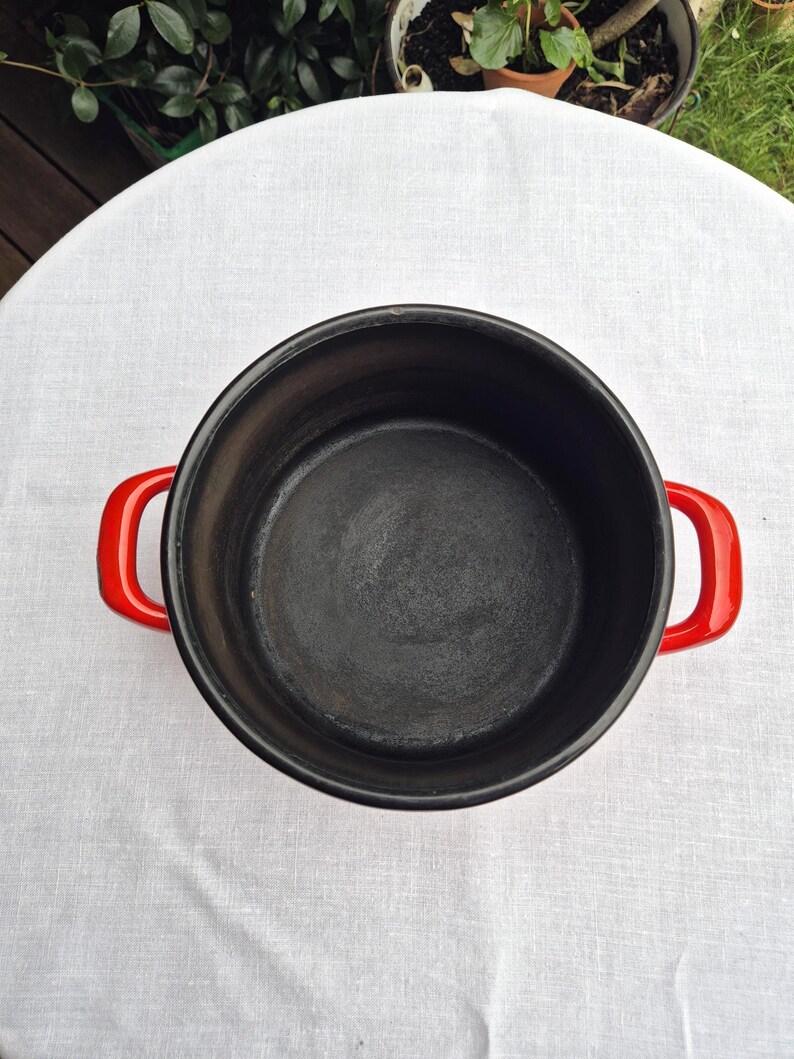 May include: A red enameled cast iron pot with black interior and two red handles. The pot is empty and sitting on a white tablecloth.