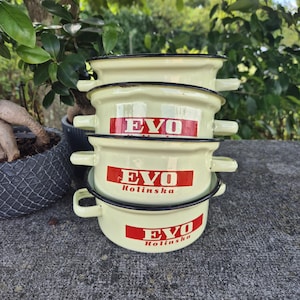 May include: A stack of four cream-colored enamelware bowls with black rims and handles. Each bowl features the red text "EVO Kolinska" on the front. The bowls are stacked on a textured gray surface.