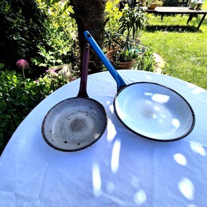 May include: Two vintage enamelware pans with long handles. One pan has a white interior and a blue handle, the other has a gray interior and a purple handle. Both pans have black rims and show signs of wear.