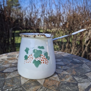 May include: White enamel pitcher with a black handle and a grape design. The pitcher has a worn, rusty rim.