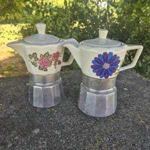 May include: Two stovetop espresso makers with white ceramic upper chambers and metallic lower bases. One features a pink rose floral pattern, while the other displays a large blue daisy-like flower. Both have matching white lids and handles.