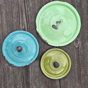 Puede incluir: Tres tapas vintage de esmalte en tonos turquesa, verde y chartreuse. Cada tapa tiene un pomo central y muestra signos de desgaste, con pintura desconchada en los bordes. Las tapas están dispuestas sobre una superficie de madera envejecida.
