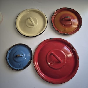 May include: Four vintage enamelware lids in different colors: white, orange, red, and blue. Each lid has a metal handle and a slightly worn appearance.