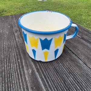 Puede incluir: Taza de esmalte blanco con borde y asa azules. La taza presenta un patrón de formas geométricas azules y amarillas. La taza está sobre una superficie de madera desgastada.