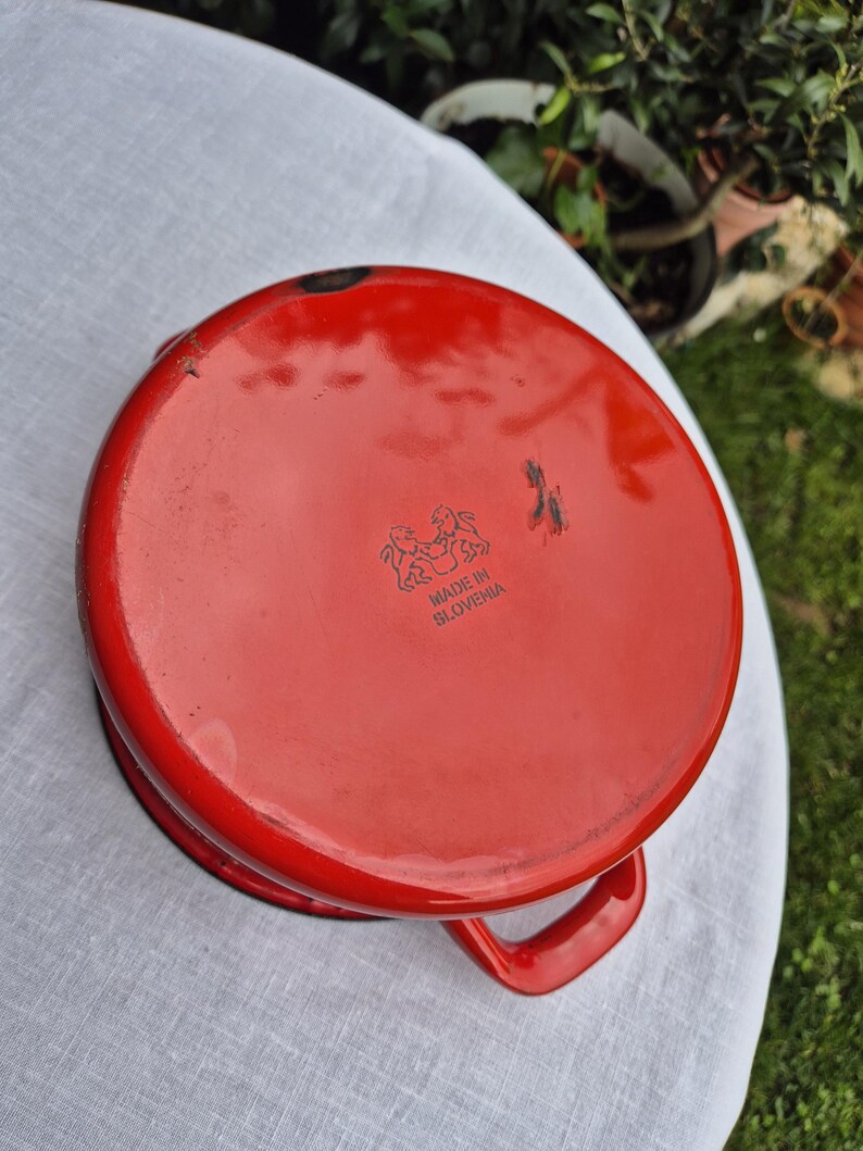 May include: Red enamel cast iron pot with a black handle and the words "Made in Slovenia" printed on the bottom.