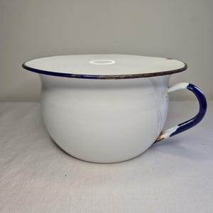 May include: White enamel chamber pot with a blue rim and handle. The antique pot has a rounded shape and a lid. The handle is a curved design with a blue and white color scheme. The rim has a dark brown edge.