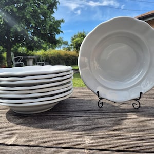 May include: A stack of white ceramic plates with scalloped edges, and one plate on a black metal stand. The plates are on a weathered wooden table outdoors, with a blurred background of trees and a house.