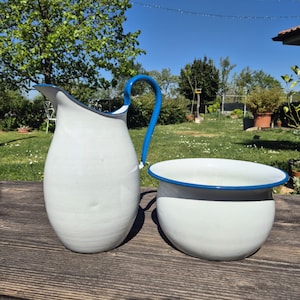 May include: A white enamel pitcher with a blue handle and rim, next to a matching bowl. The pitcher has a curved handle and a spout. The bowl is round with a blue rim. Both items are on a wooden surface.