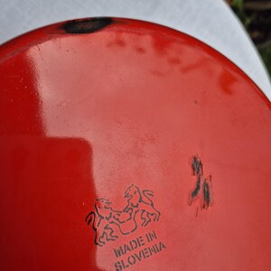 May include: Red enamelware pot with a black rim and a black logo that says "Made in Slovenia" with two lions facing each other.